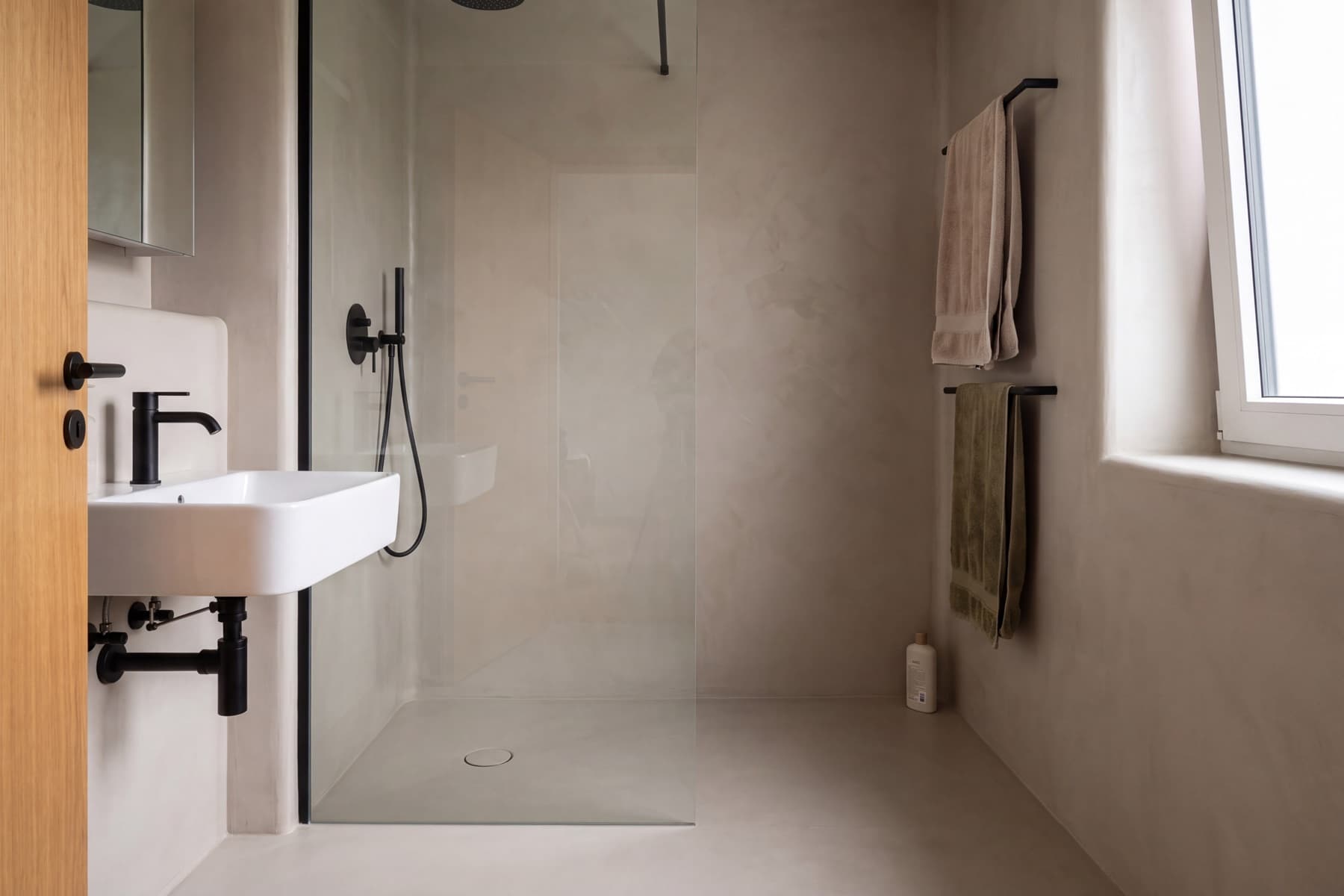 Bad ohne Fliesen: Wände und Boden in fugenlosem, warm graubeigem Spachtelbelag, dazu ein wandhängendes Waschbecken mit schwarzer Armatur und eine Walk-in-Dusche mit flacher heller Duschwanne hinter einer Glasabtrennung.