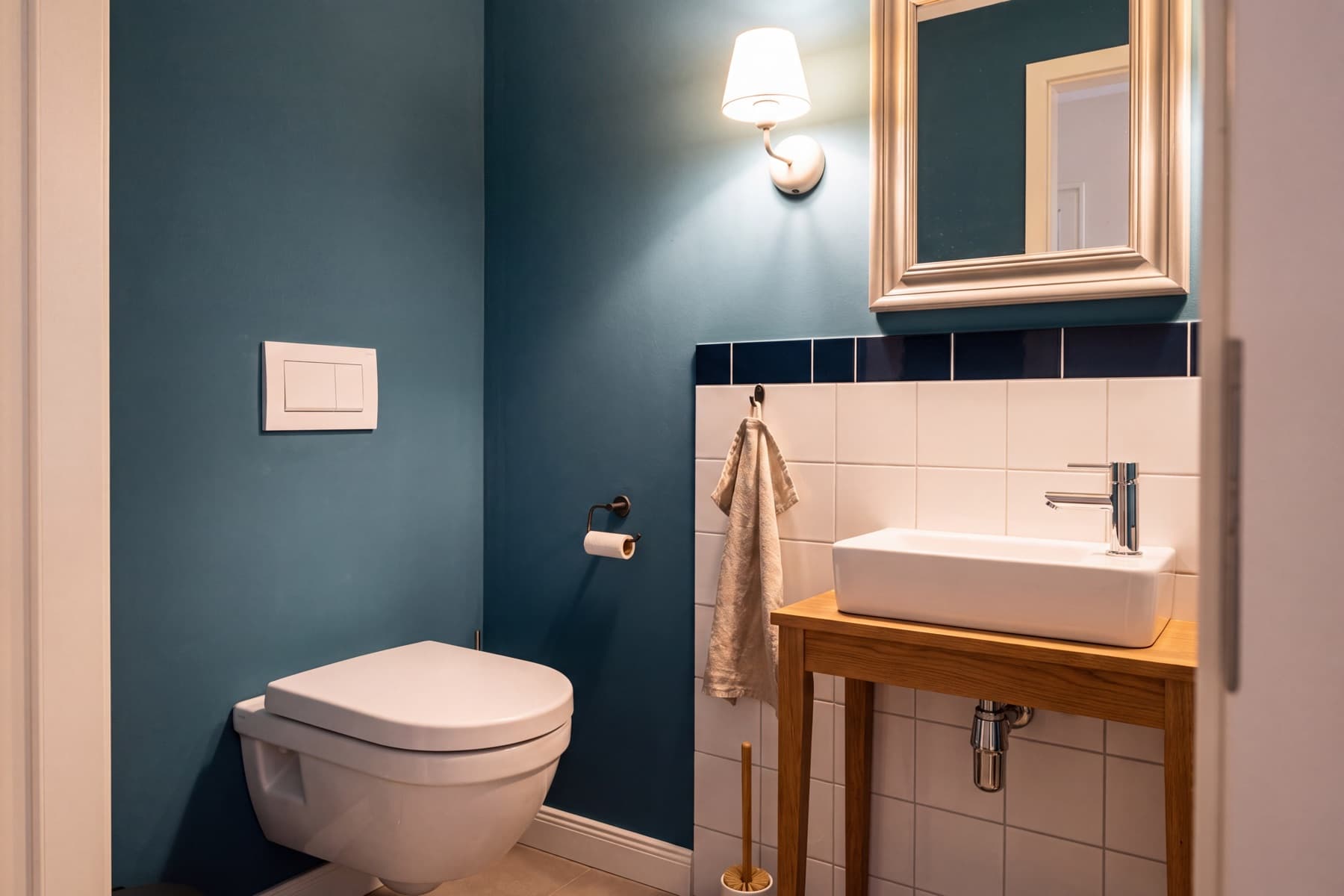 Fertiges Gäste-WC ohne Fenster: petrolblaue Wand mit weißem wandhängendem WC und schlichter weißer Betätigungsplatte, rechts ein weißes Aufsatzbecken auf einer Eichenkonsole, darüber ein gerahmter Spiegel und eine Wandleuchte, weiße Fliesen bis zur halben Höhe