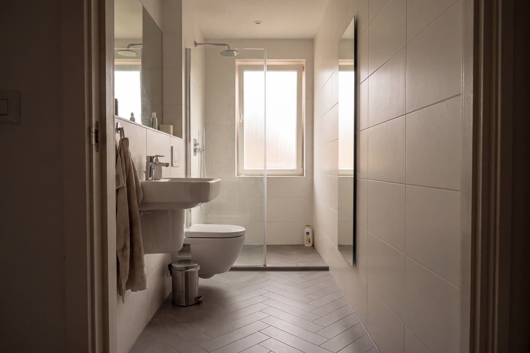 Schmales, langes Bad vom Türrahmen der Länge nach gesehen: links Waschtisch und wandhängendes WC in einer Linie, rechts eine geflieste Wand, am Ende unter dem Fenster eine bodengleiche Dusche hinter einer Festverglasung.