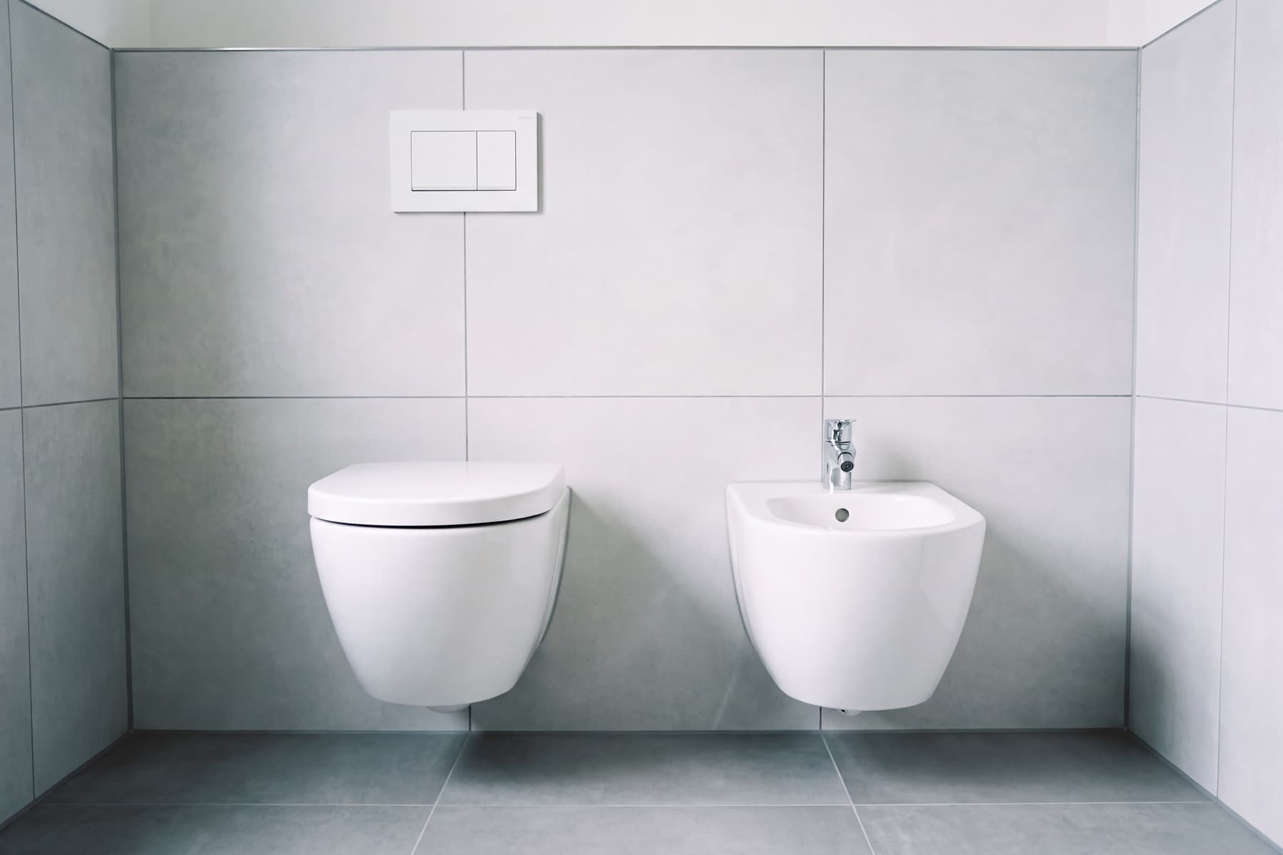 Fertig montierte Sanitärobjekte an einer Wand aus grauen Großformatfliesen: ein weißes wandhängendes WC und daneben ein weißes Wandbidet, beide frei über dem Boden, darüber eine schlichte weiße Betätigungsplatte