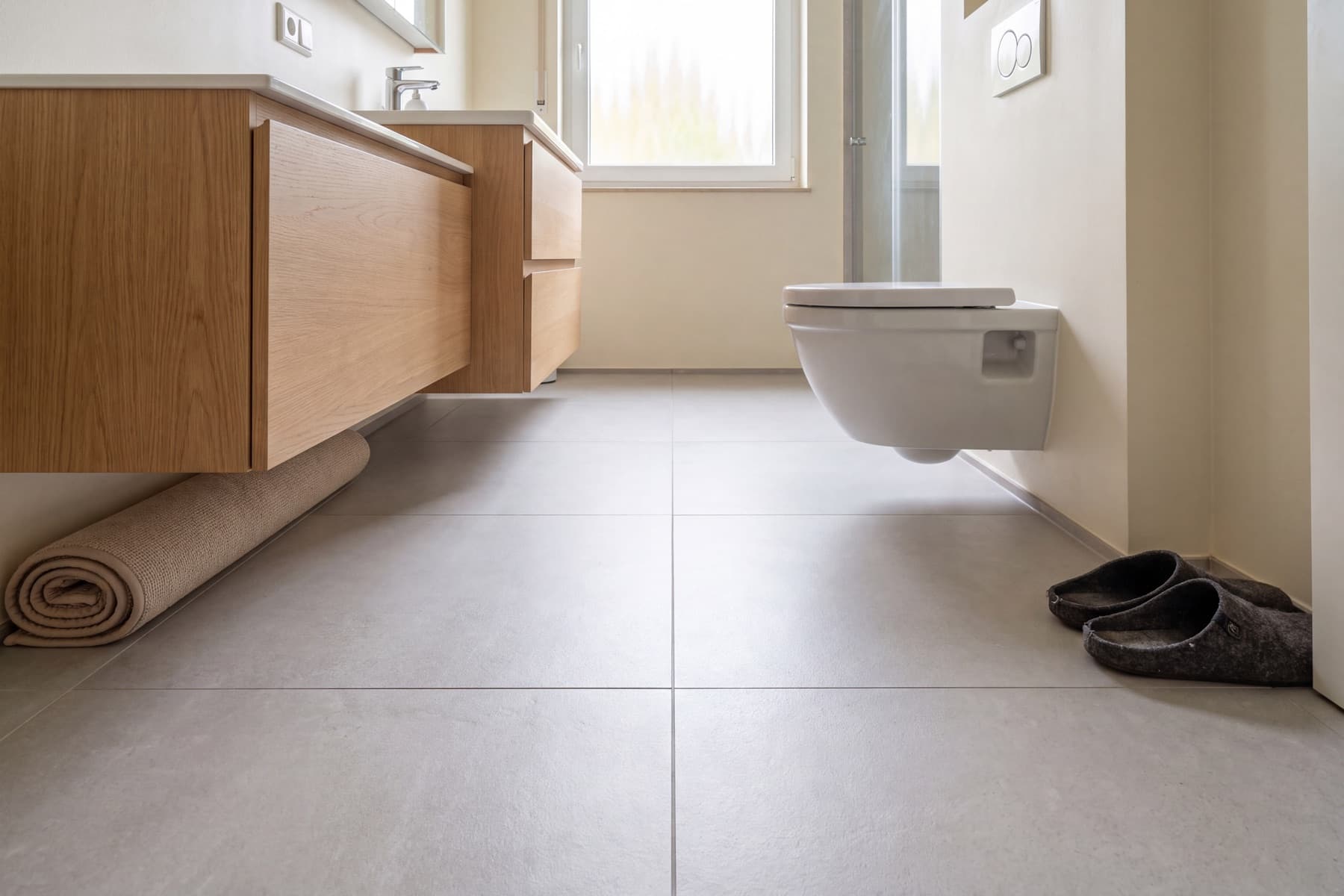Flache Aufnahme knapp über dem Boden: der graue Fliesenboden läuft ohne Unterbrechung unter einem wandhängenden WC und einem wandhängenden Waschtischunterschrank aus Eiche hindurch bis zur gegenüberliegenden Fensterwand; links lehnt ein zusammengerollter Badvorleger, rechts stehen Filzpantoffeln.