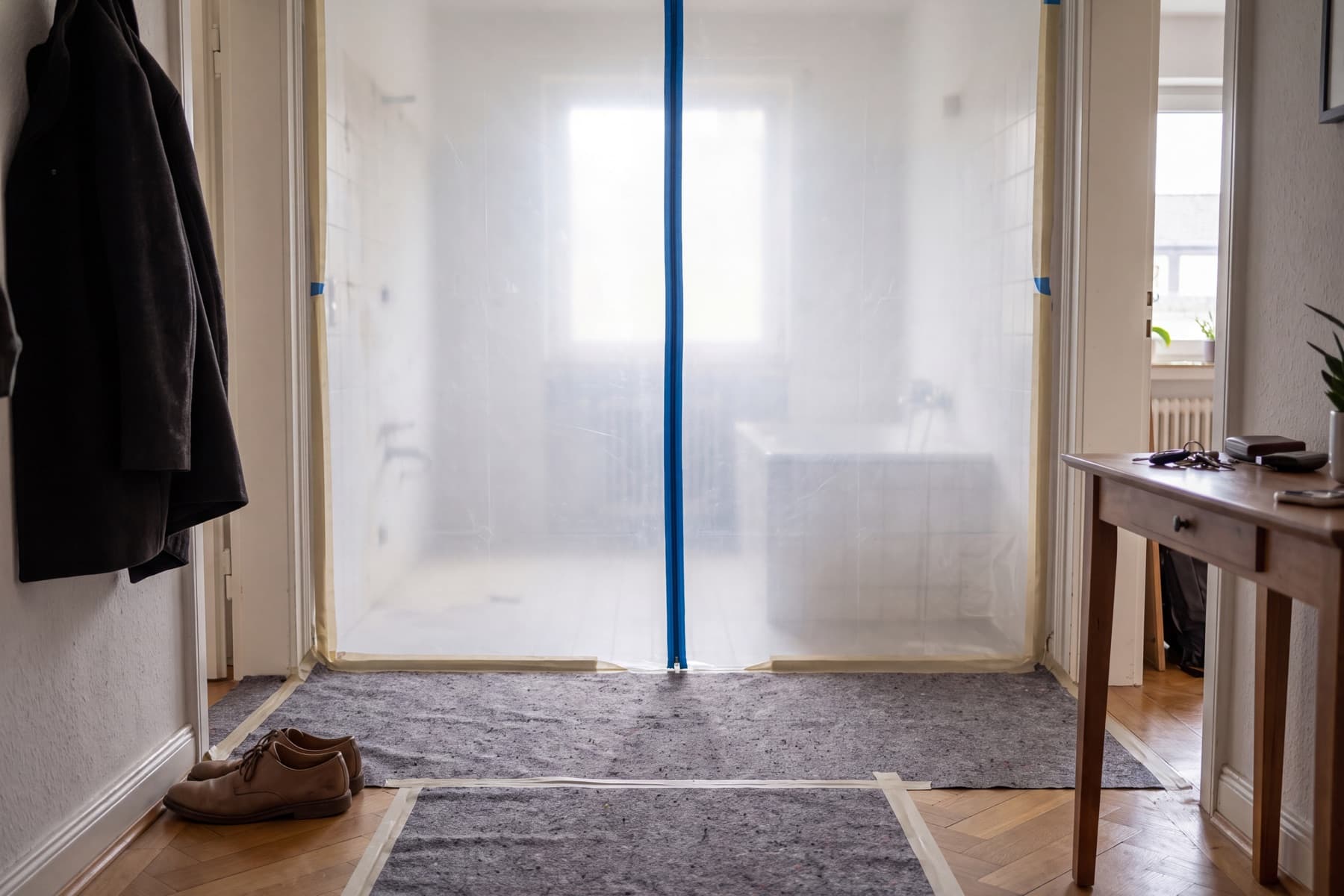 Staubschutzwand aus durchscheinender Folie mit Reißverschluss vor der Badtür im Flur, davor ein abgeklebtes Abdeckvlies auf dem Parkett, dahinter schemenhaft das ausgeräumte alte Bad mit Wanne und Heizkörper.