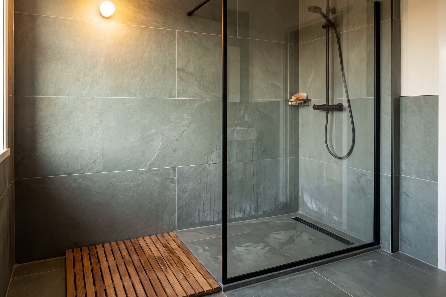 Walk-in-Dusche mit einer feststehenden Scheibe in schmalem schwarzem Rahmen und offenem Zugang, schräg von der Seite gesehen: grüngraue Fliesen in Schieferoptik, mattschwarze Armaturen und eine Rinne an der Wandkante.