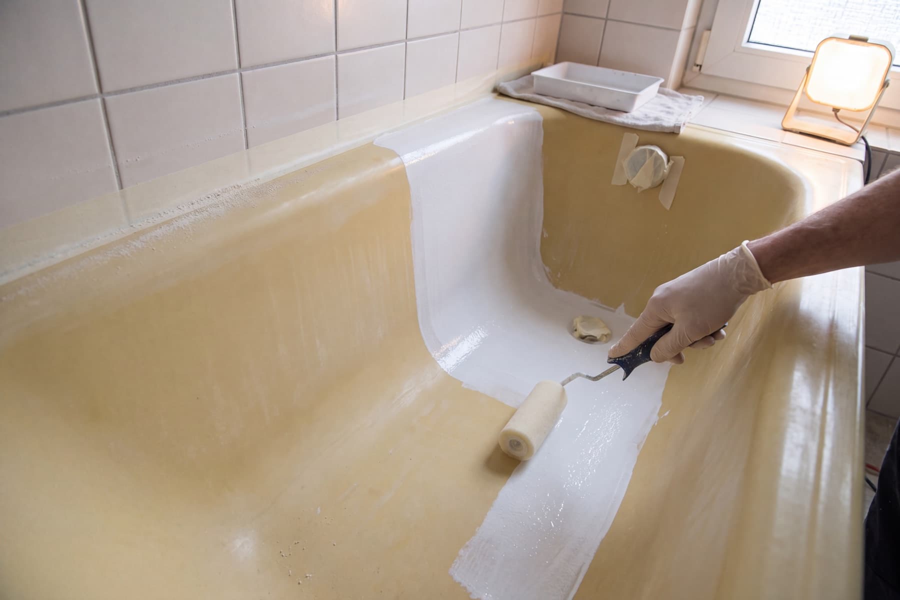 Eine Hand im Latexhandschuh rollt mit einem Farbroller weiße Beschichtung über eine stark vergilbte Badewanne; die frisch beschichtete Bahn ist deckend weiß, die unbehandelte Fläche daneben gelb und glänzend, Ablauf und Überlauf sind mit Kreppband abgeklebt.